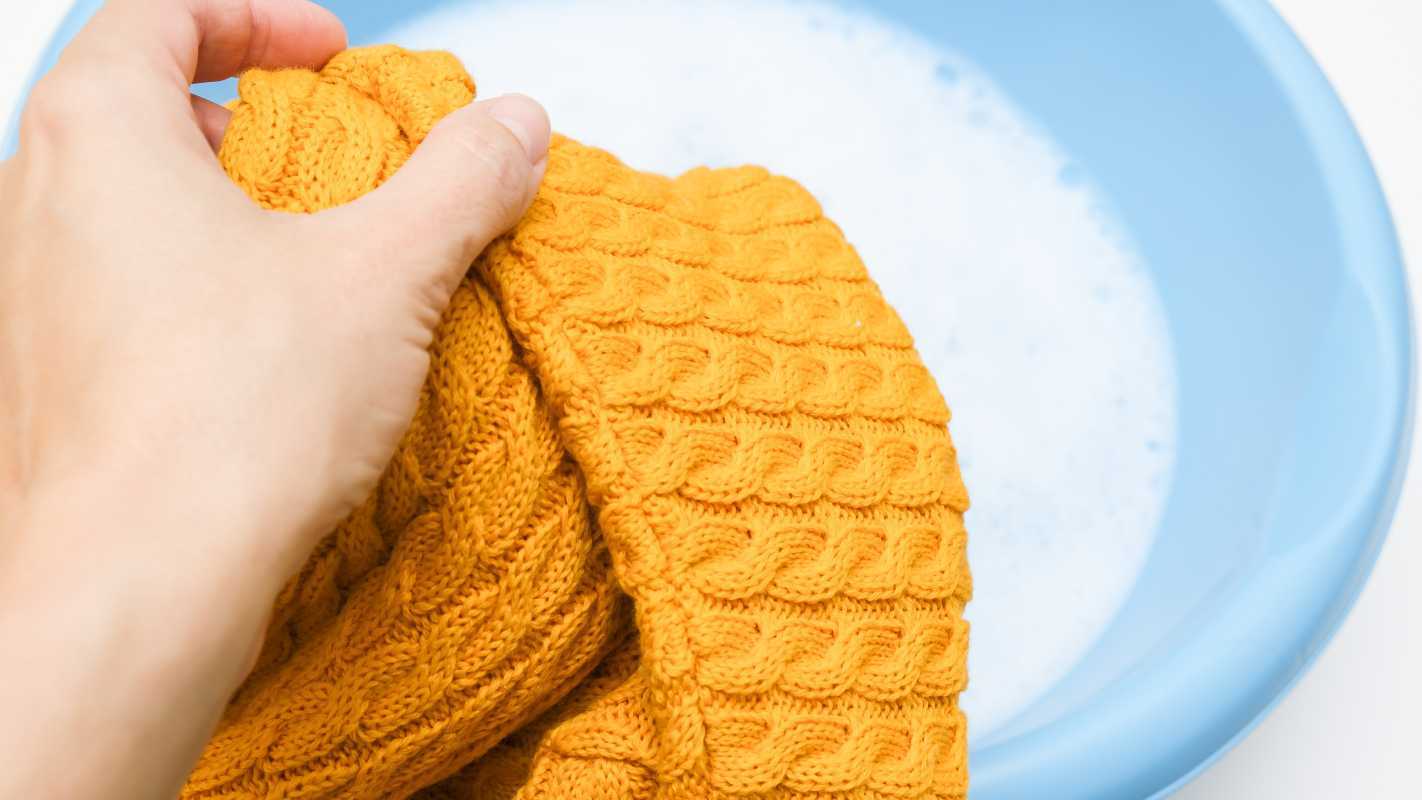 Mujer lavando a mano una manta de crochet amarilla en un recipiente con agua jabonosa