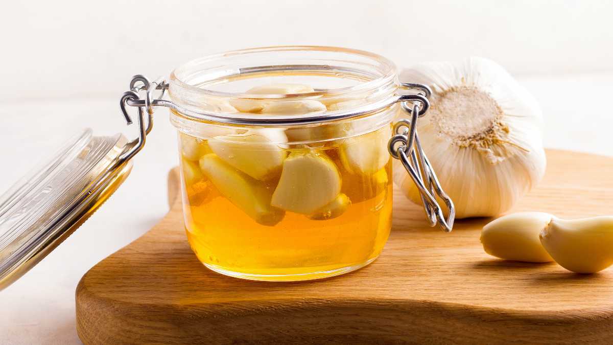 Toples kaca lebar berisi madu dan bawang putih, yang memiliki manfaat kesehatan, di atas talenan kayu