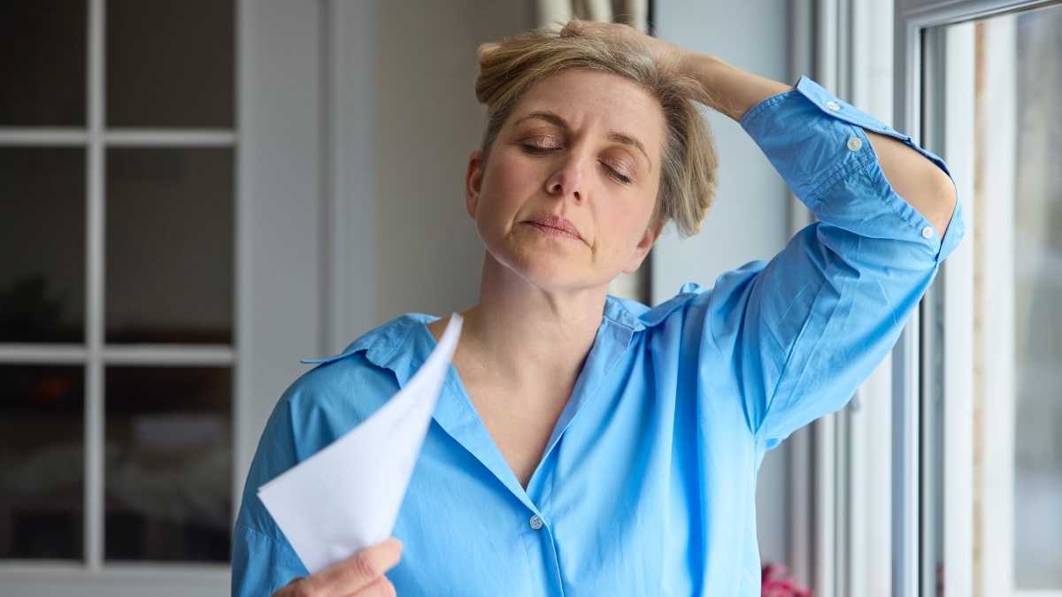 Een blonde vrouw in een blauw shirt die haar haar omhoog houdt en zichzelf uitwaaiert met papier vanwege een opvlieger