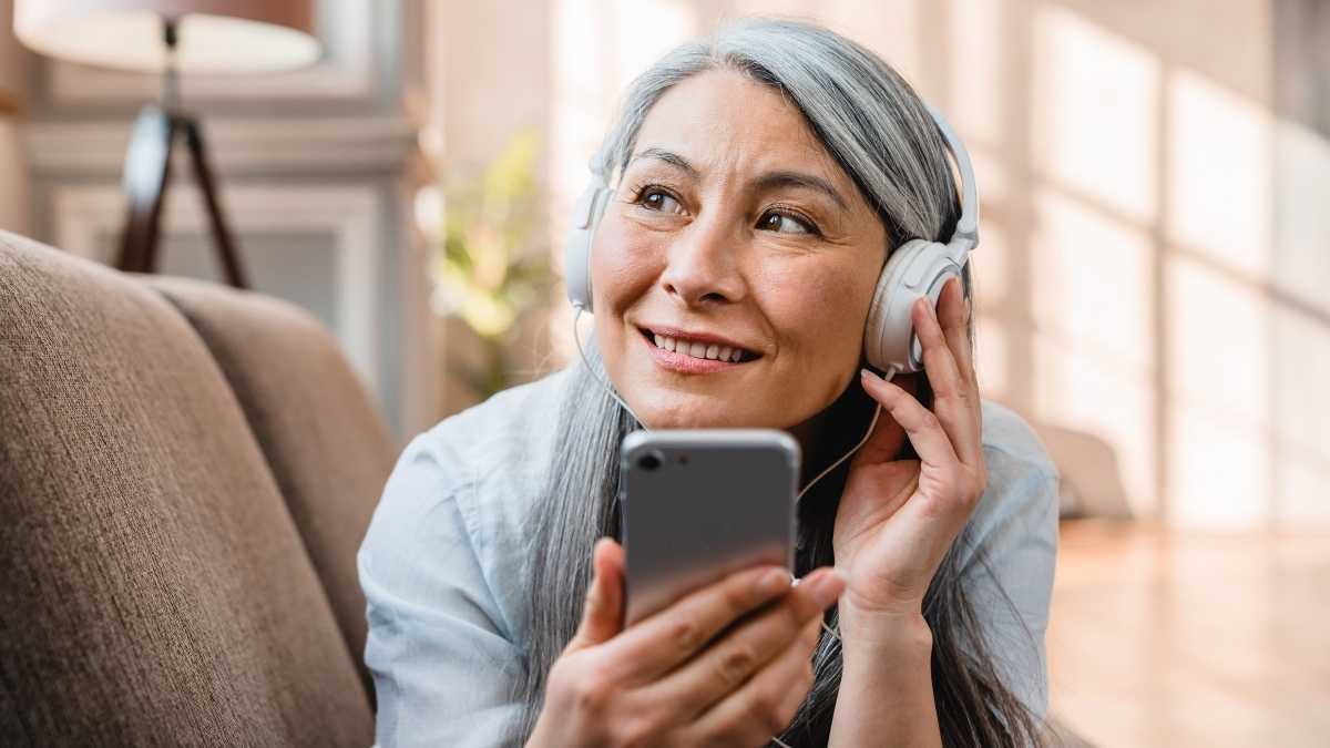 Žena s šedými vlasy se sluchátky držící telefon a poslouchá audioknihu
