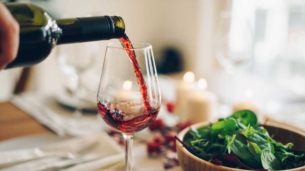 En flaske rødvin hældes i et klart glas ved siden af ​​en skål spinat