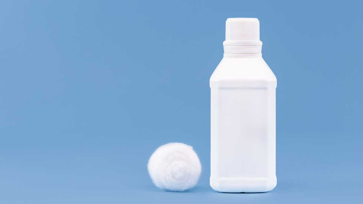 En vit flaska väteperoxid bredvid en vit bomullstuss på en blå bakgrund