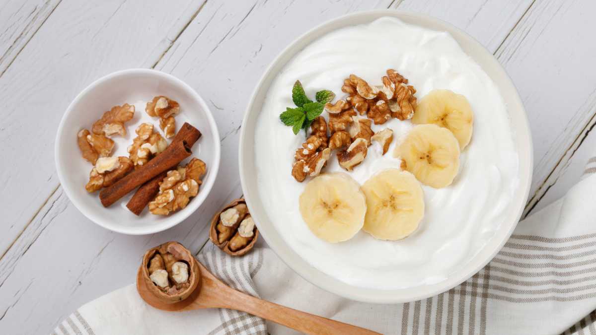 Een kom yoghurt, een van de beste huismiddeltjes tegen bacteriële vaginose, gegarneerd met gesneden bananen en walnoten op een houten tafel